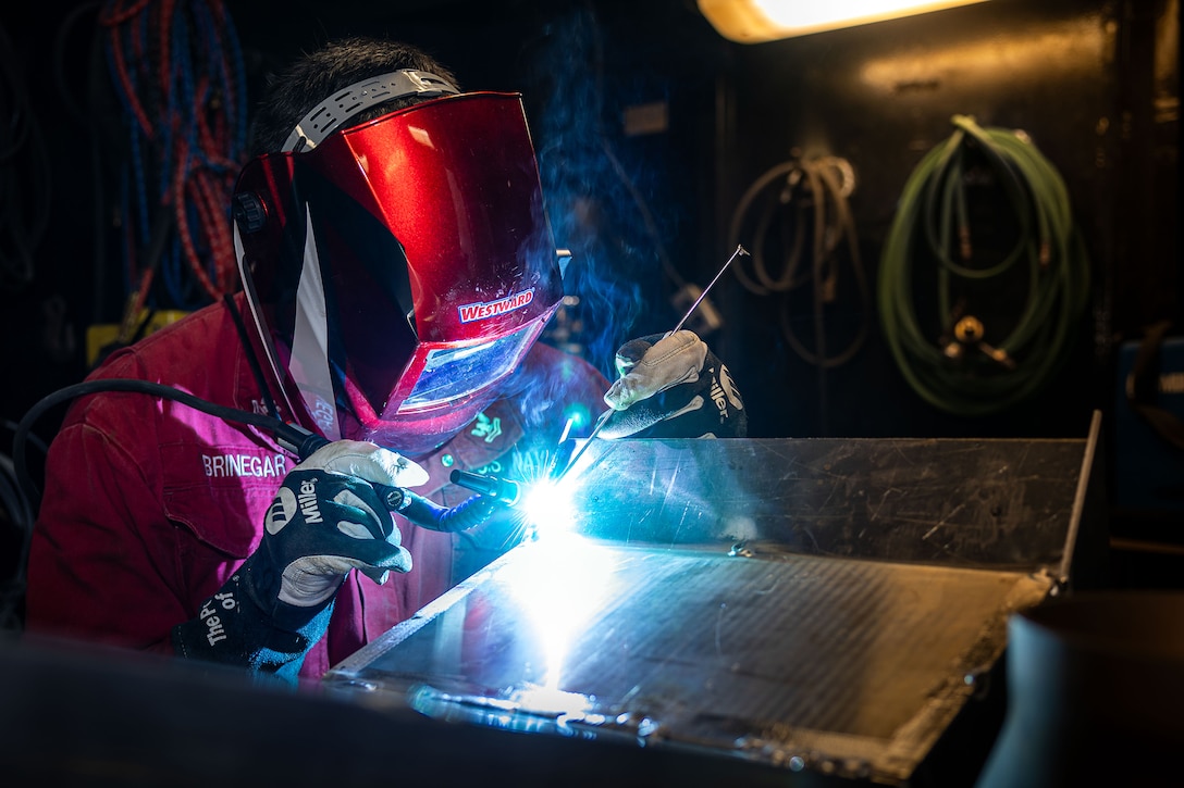 A sailor wearing a welding helmet and gloves welds a piece of equipment in a repair shop creating a bright, white flame.