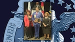 Two women and one man stand in front of flags. The man is holding an award plaque