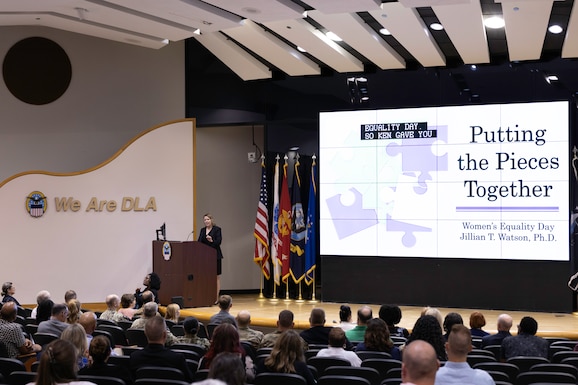 A woman in a black dress stands at a podium delivering a lecture on Equality with a slideshow next to her in a large auditorium.