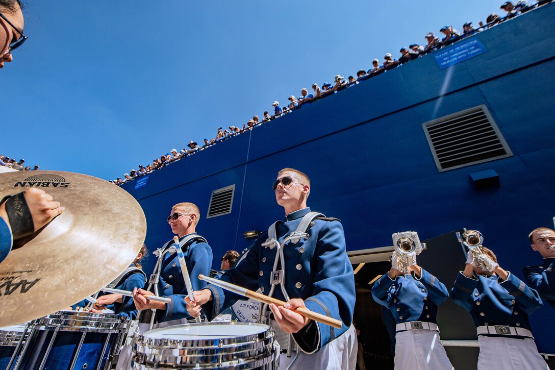 Dozens of spectators line up against a stadium and look down as a band performs.