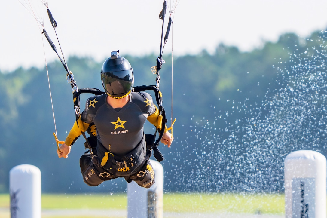 A soldier wearing a helmet with a parachute performs while descending as water splashes from the right.