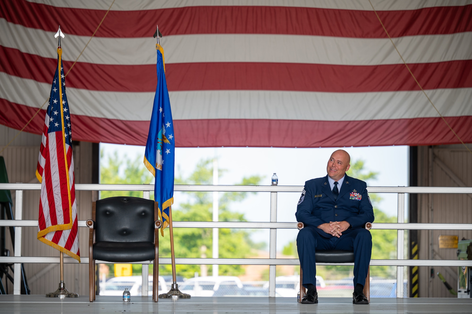 A man in a U.S. Air Force dress uniform sits in a chair on a stage. To his right, an empty chair flanked by an Air Force flag and American flag. In the background, the three bottom stripes of a large American flag are visible.
