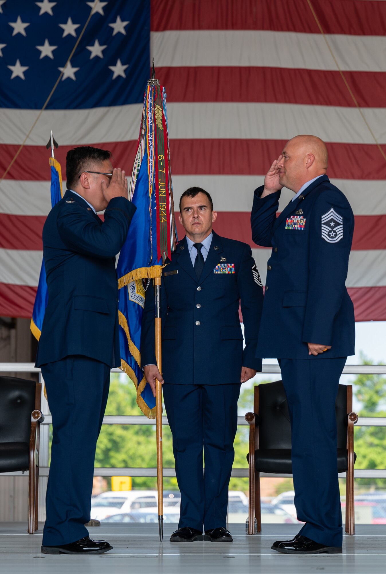 Three men in U.S. Air Force dress uniforms stand on a stage. The man at left and man at right are facing one another, saluting. The man in the middle is facing the viewer, holding a ceremonial flag.