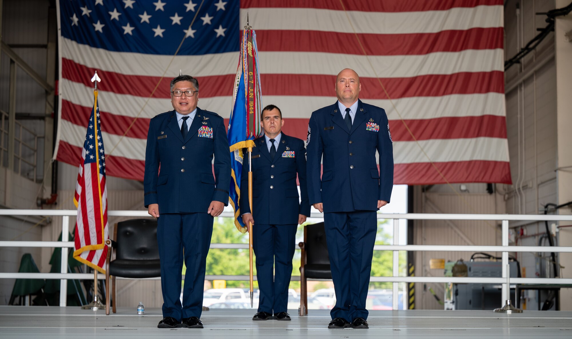 Three men in U.S. Air Force dress uniforms stand on a stage. The middle is holding a flag. The other two are standing with their hands at their sides. None are looking at the camera. In the background, a giant American flag hangs from ceiling.