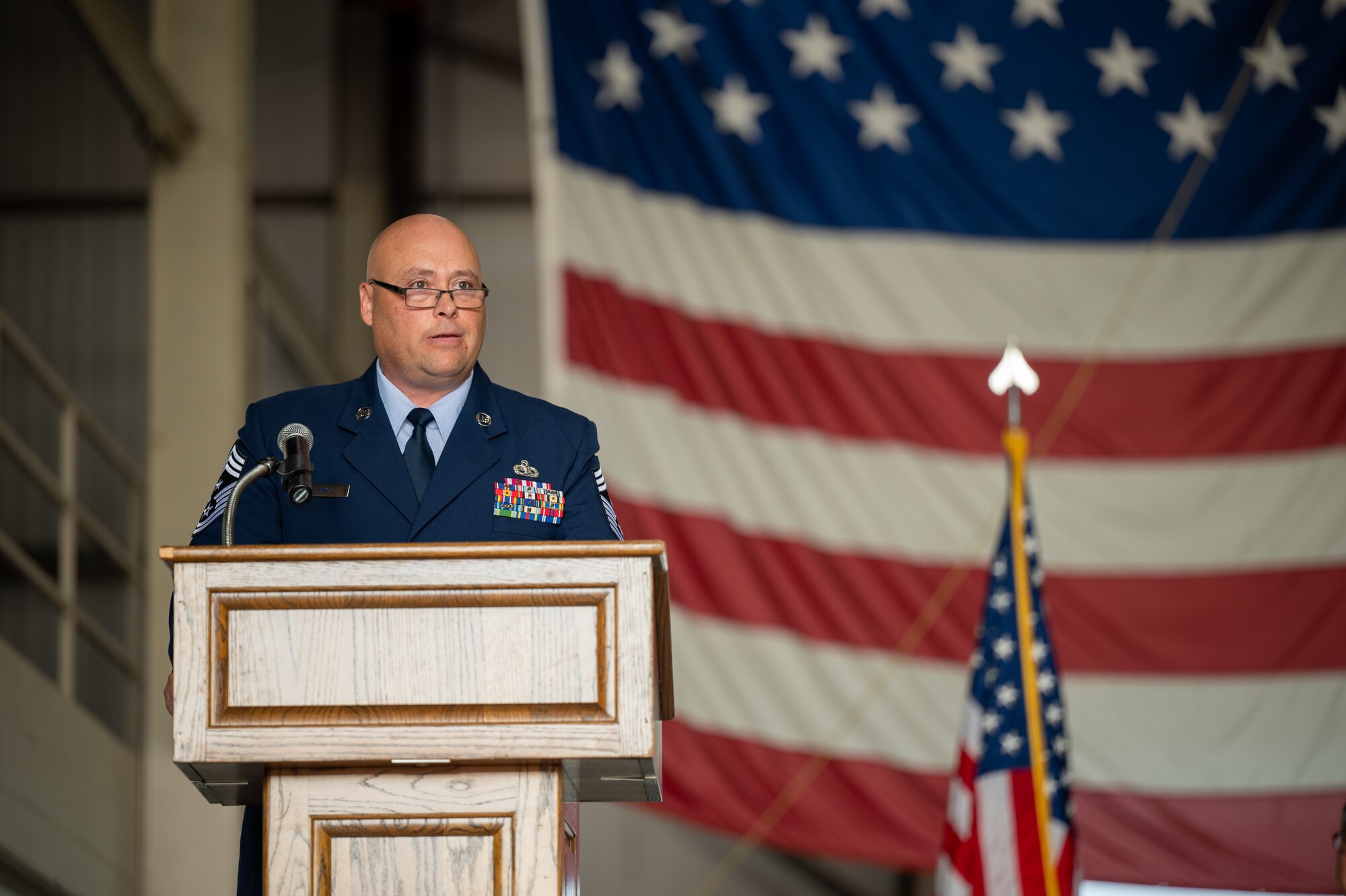 A man in a U.S. Air Force dress uniform stands behind a lectern. He is wearing glasses. In the background, two American flags. One partially visible and giant, the other smaller and on an indoor flagpole.