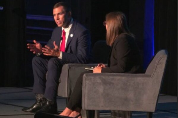 A man is seated in a chair that is turned slightly toward a woman seated next to him. The man is gesturing with his hands and appears to be speaking.