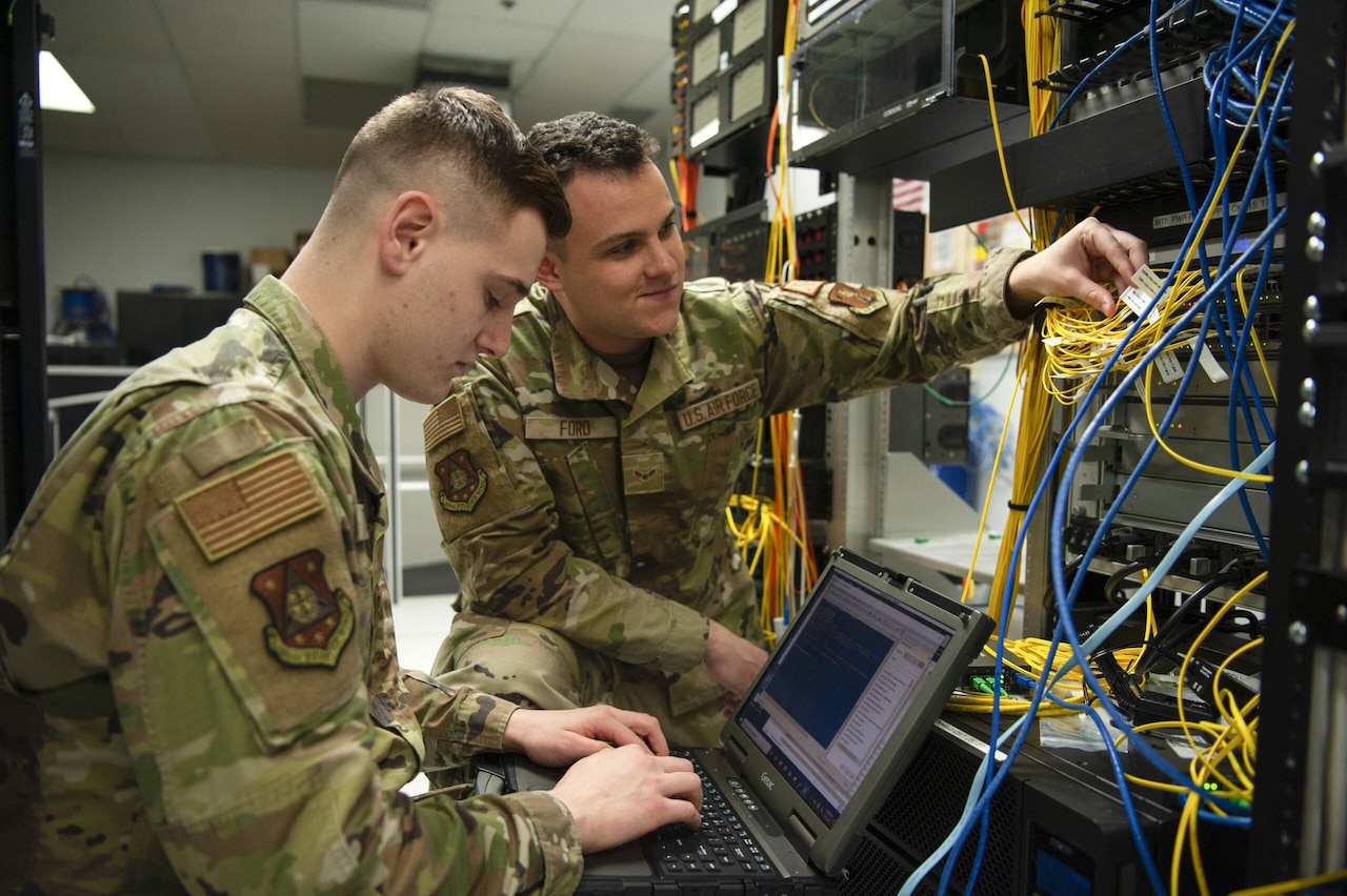 A service member manages server cables while another service member types on a laptop computer.