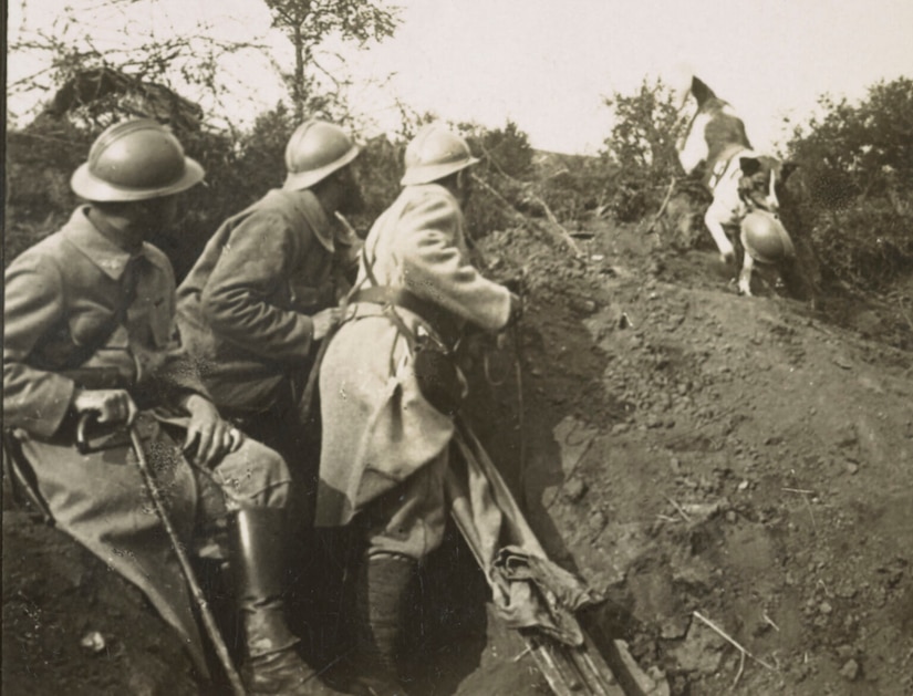 A dog holding a helmet in its mouth trots down a small hill toward three men.