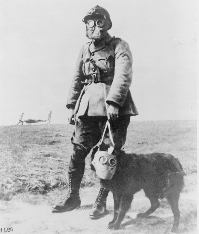 A soldier holds the leash of a dog. Both wear gas masks.