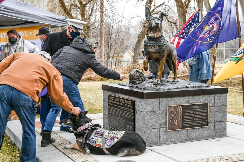 People look at a bronze statue of a dog and a helmet. A real service dog sits in front of it.