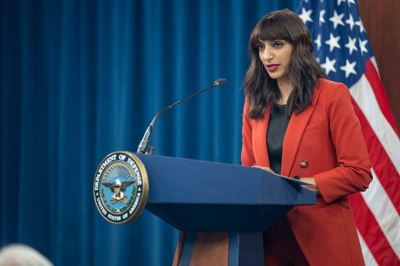 A woman is standing at a lectern speaking into a microphone while looking off screen to the left. The Defense Department logo is on the front of the lectern.