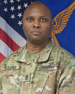 Army Soldier in green combat uniform (camouflage) posing for a portrait photo in front of flags.