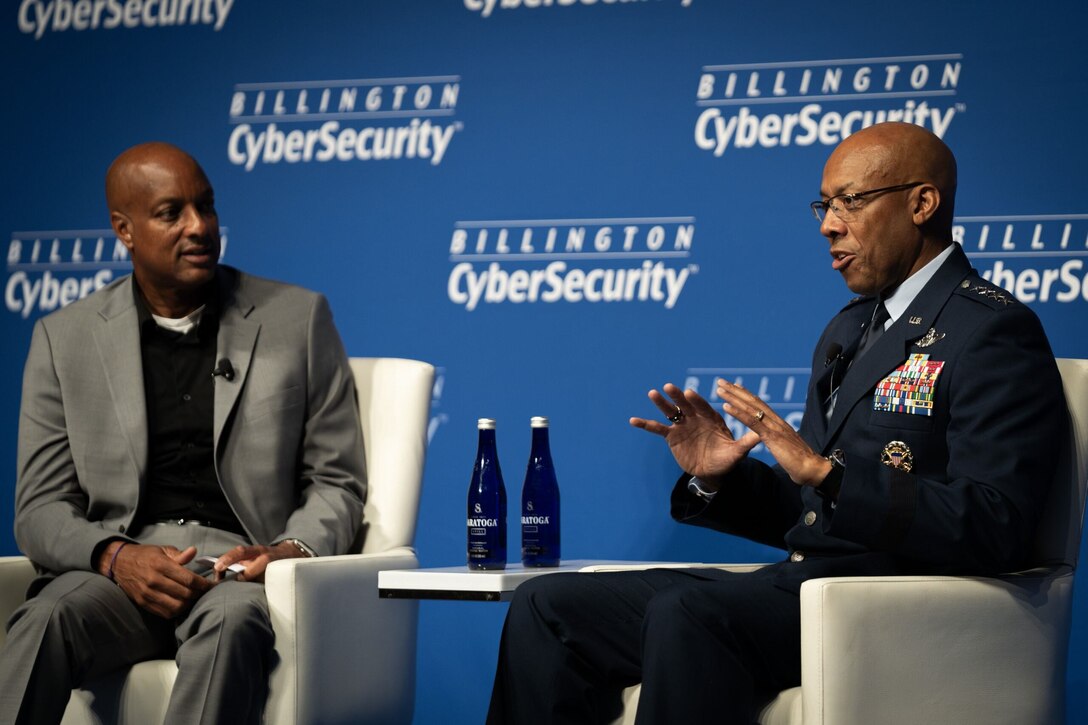 A service member sits next to a man in civilian attire on a stage.