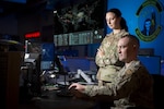 A service member stands, looking down toward a computer, while a second service member is seated at the computer.