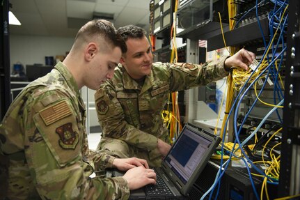 A service member manages server cables while another service member types on a laptop computer.