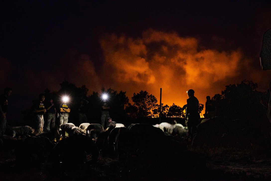 Soldiers perform pushups at night while other soldiers stand and watch. Smoke swirls above them and orange light is in the background.