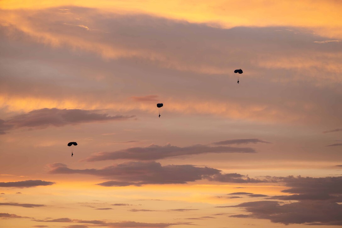 Three airmen parachute to the ground against a gray and yellow partly cloudy sky.