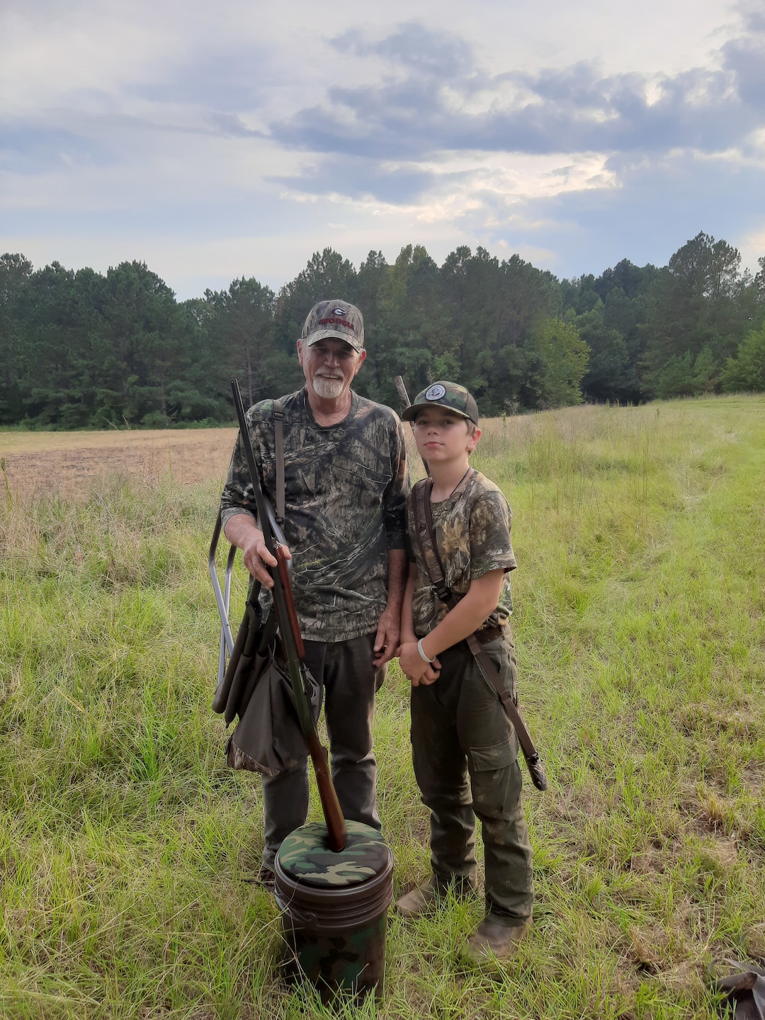 Man and child stand in field with hunting gear.