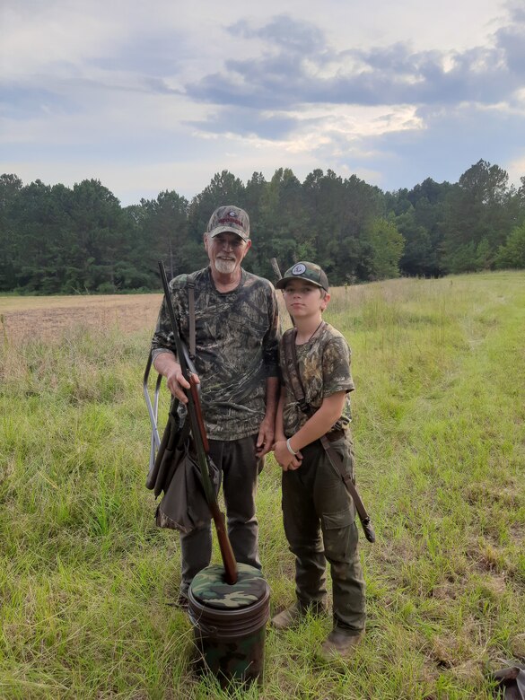 Man and child stand in field with hunting gear.