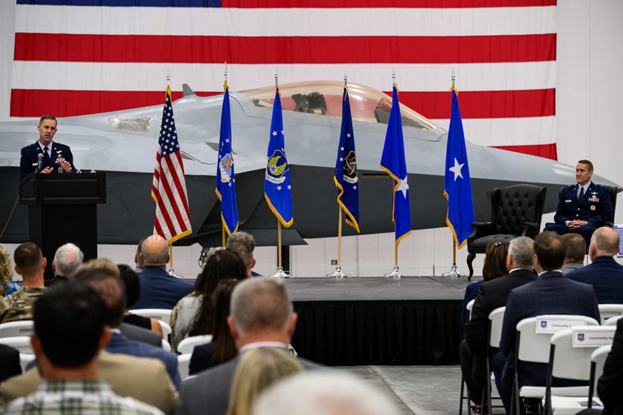 Man at lectern speaks to audience in front of a plane and six flags; second man sits on stage in chair