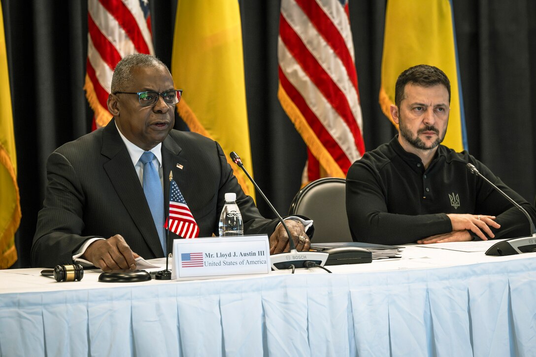 Secretary of Defense Lloyd J. Austin III speaks while seated at a white covered table next to the Ukrainian president with American and Ukrainian flags in the background.