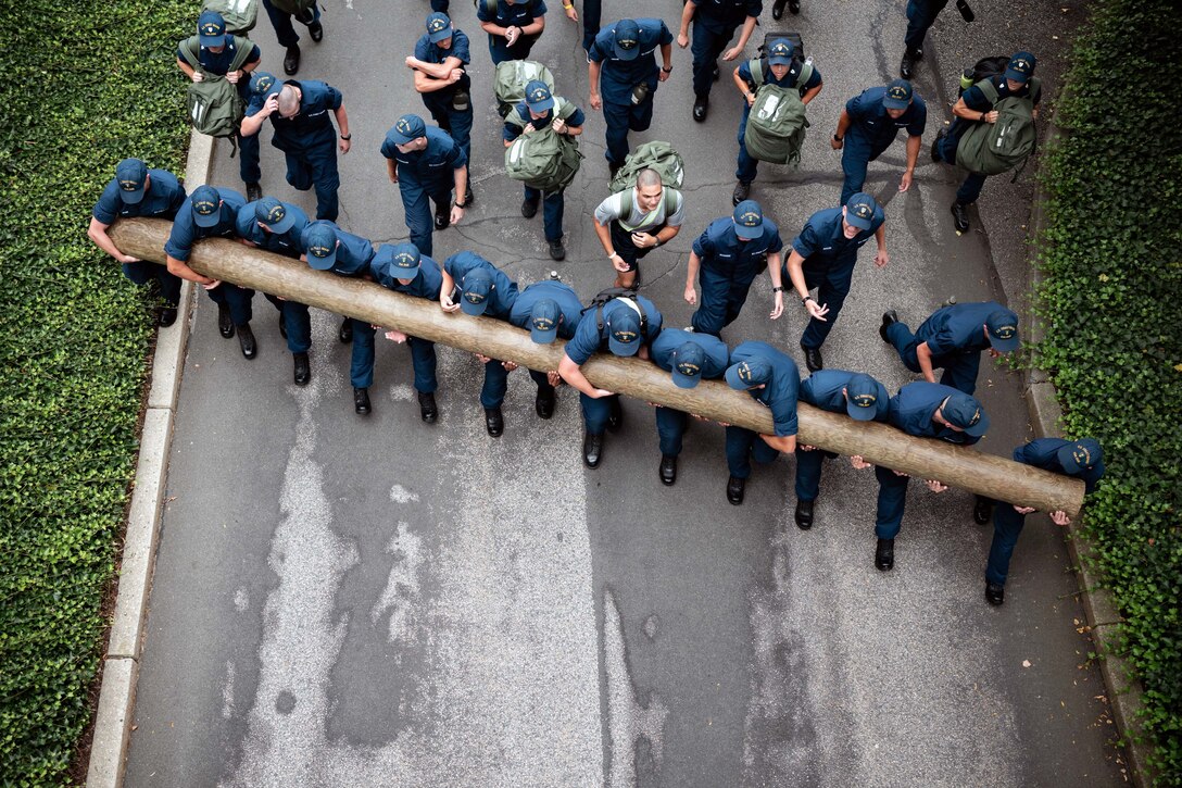 From overhead, a group of Coast Guardsmen in training uniform hold a log diagonally while walking on a street with more others following.