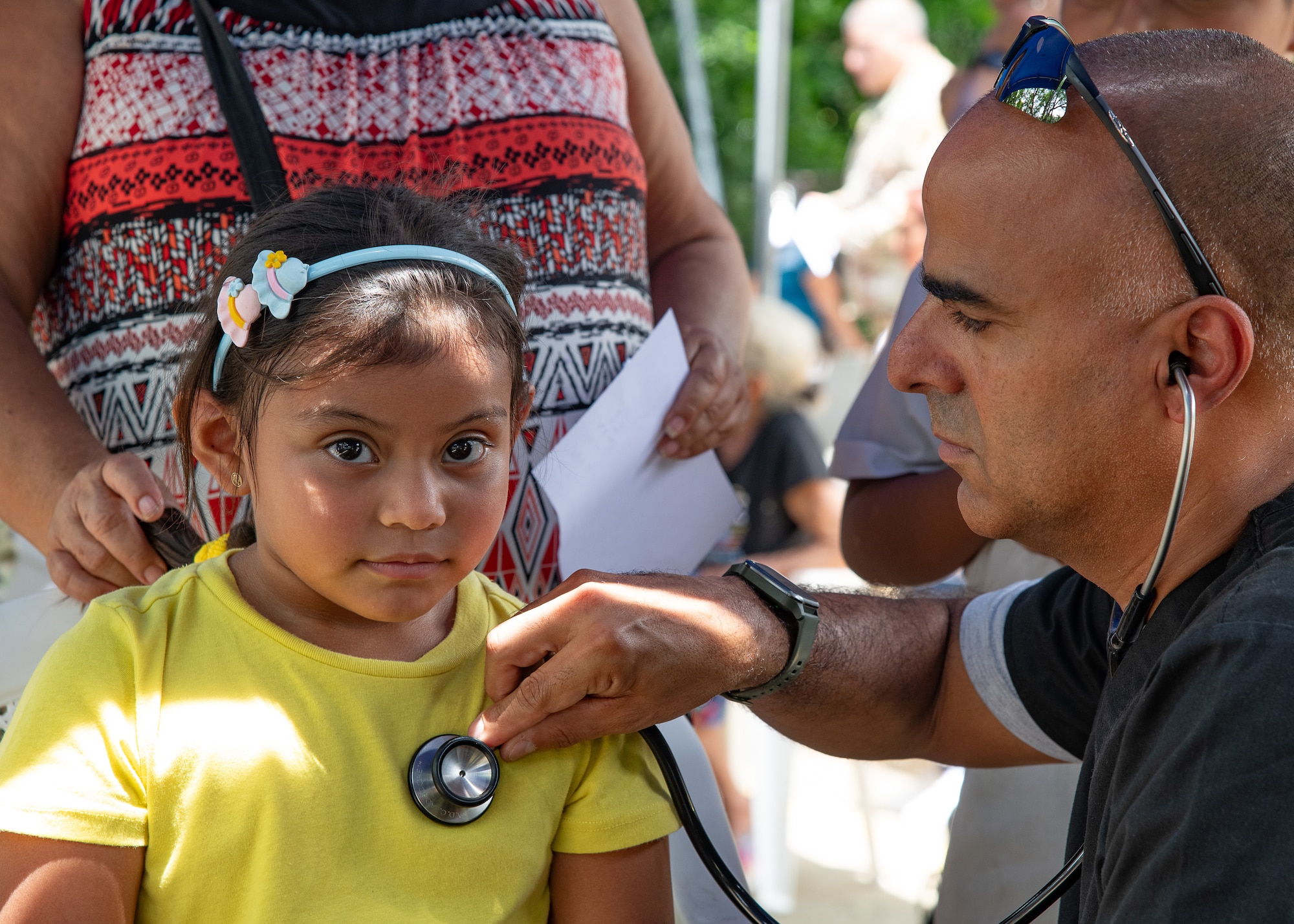 A man holds a stethoscope to a child's chest while her mother stands behind her.
