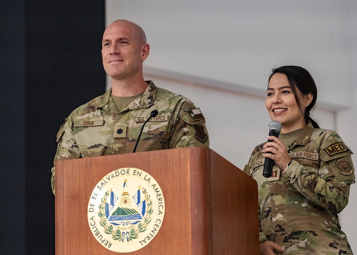 Two U.S. military members stand behind a podium with an emblem on it that reads "REPUBLICA DE EL SALVADOR EN LA AMERICA CENTRAL"