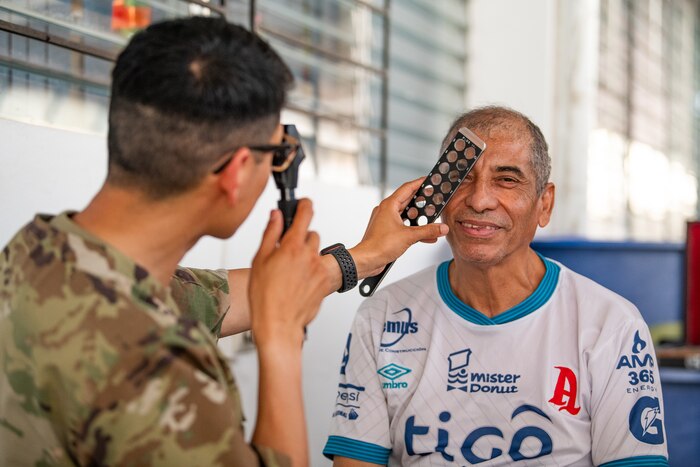 A man looks through an optometry tool while a second man in U.S. military uniform examines him.