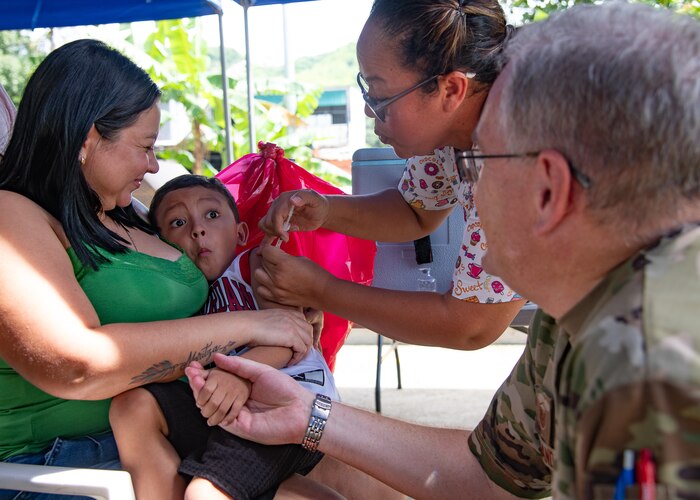 A child receives a shot while being held by a woman and holding the hand of a man in U.S. military uniform.