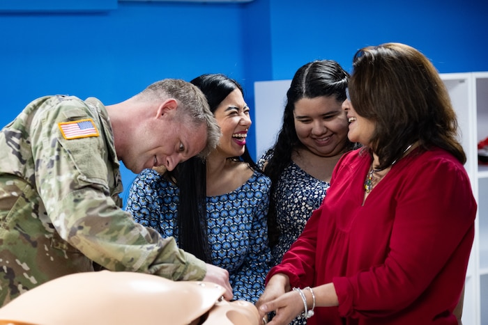 A man in U.S. military uniform manipulates a training mannequin while three women in civilian clothes laugh amongst each other.