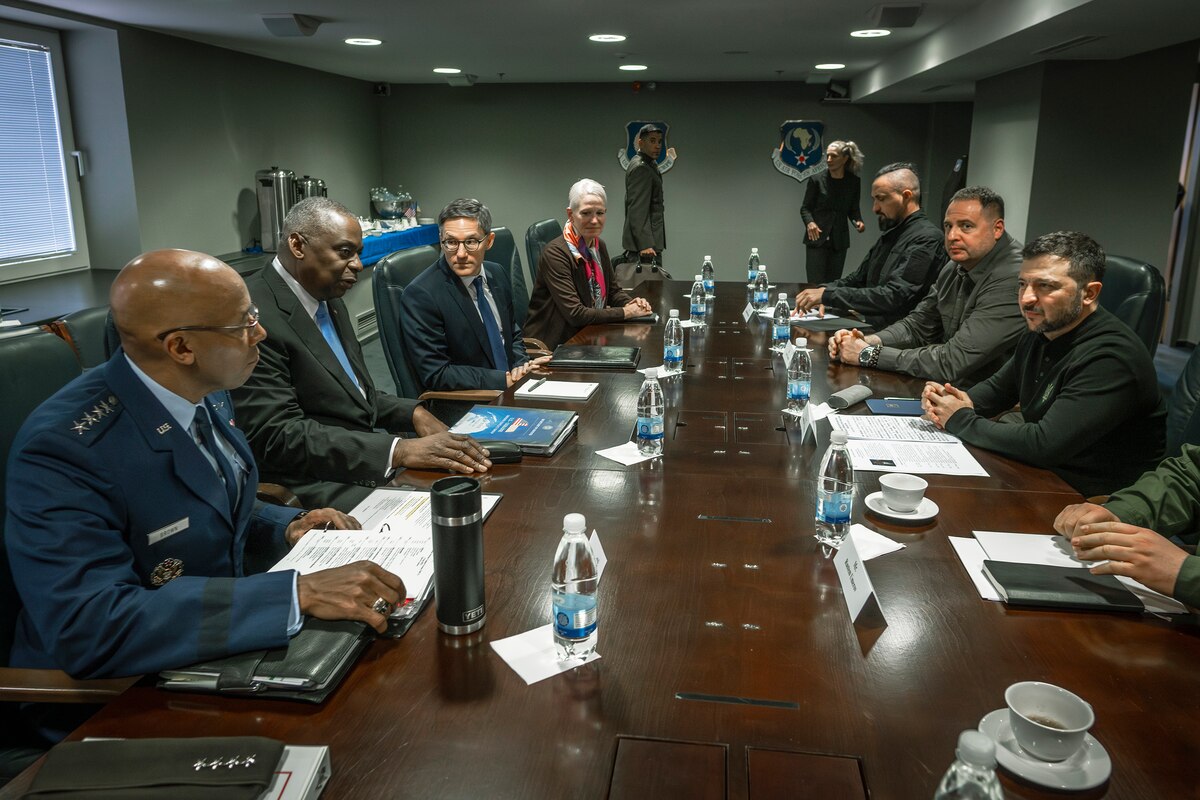 A group of eight men, most in business attire and one in a dress military uniform, sit around a large wooden table with papers and bottled water within their reach.