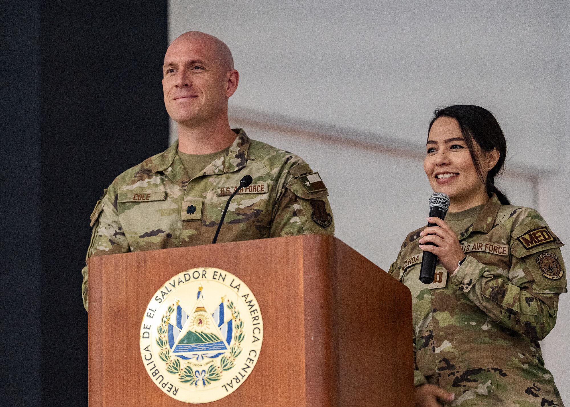 Two U.S. military members stand behind a podium with an emblem on it that reads "REPUBLICA DE EL SALVADOR EN LA AMERICA CENTRAL"