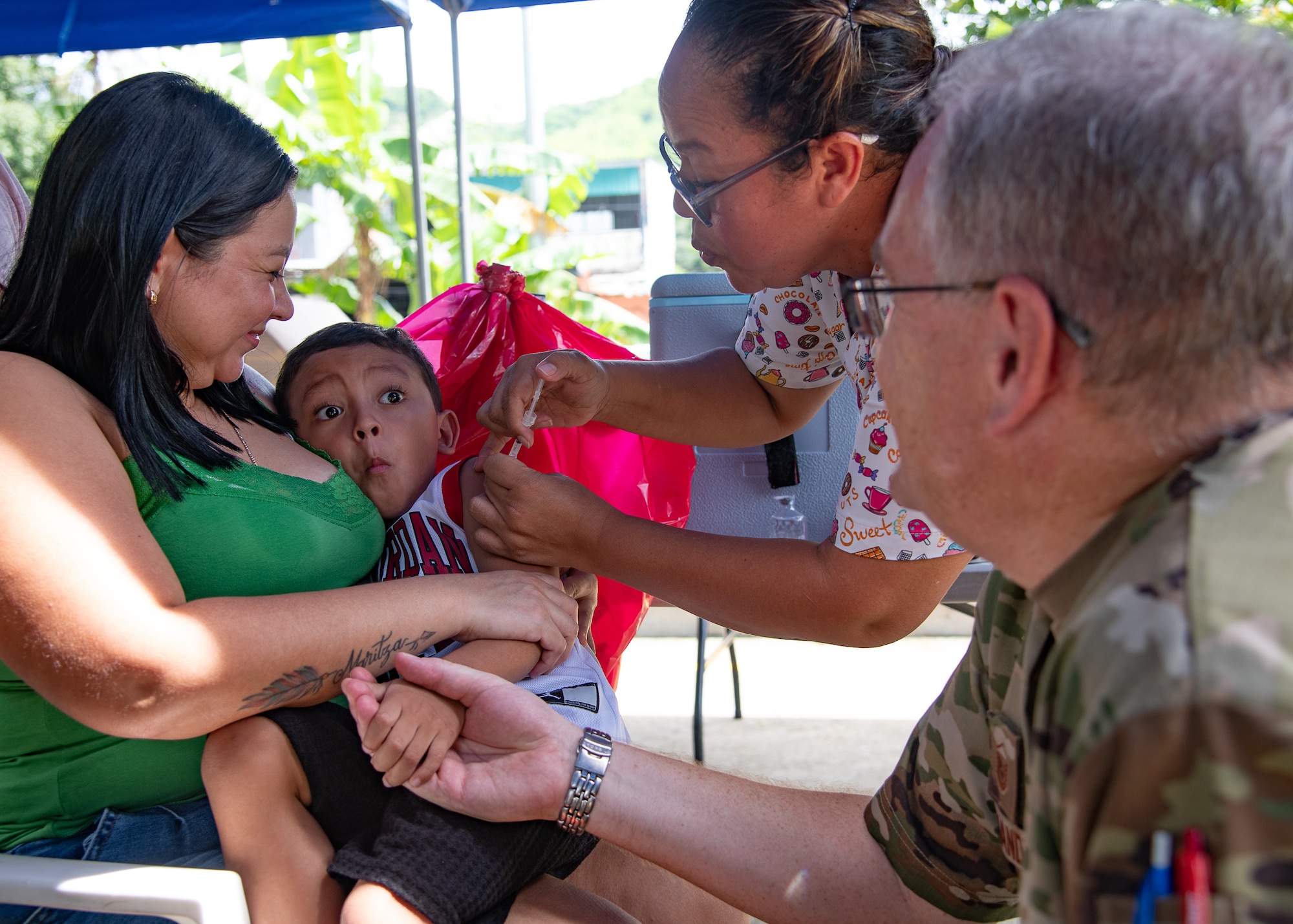 A child receives a shot while being held by a woman and holding the hand of a man in U.S. military uniform.