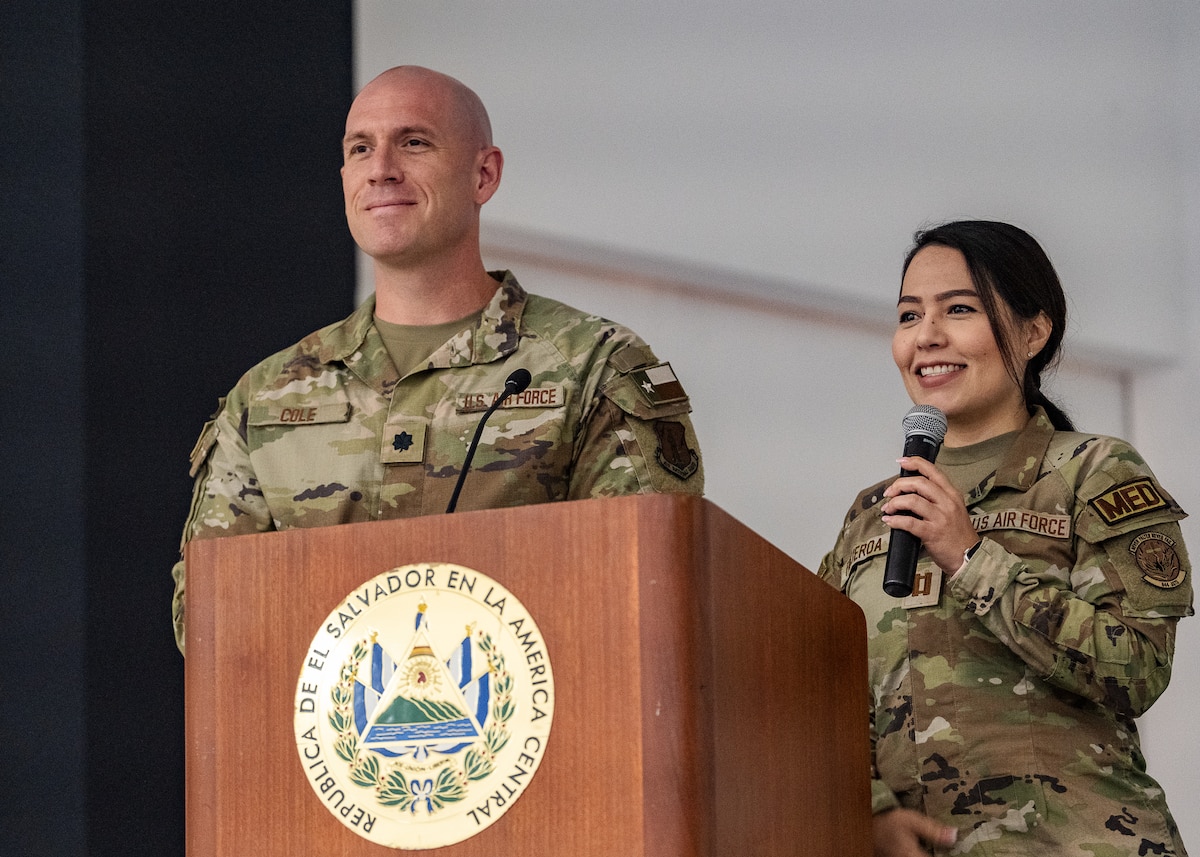 Two U.S. military members stand behind a podium with an emblem on it that reads "REPUBLICA DE EL SALVADOR EN LA AMERICA CENTRAL"