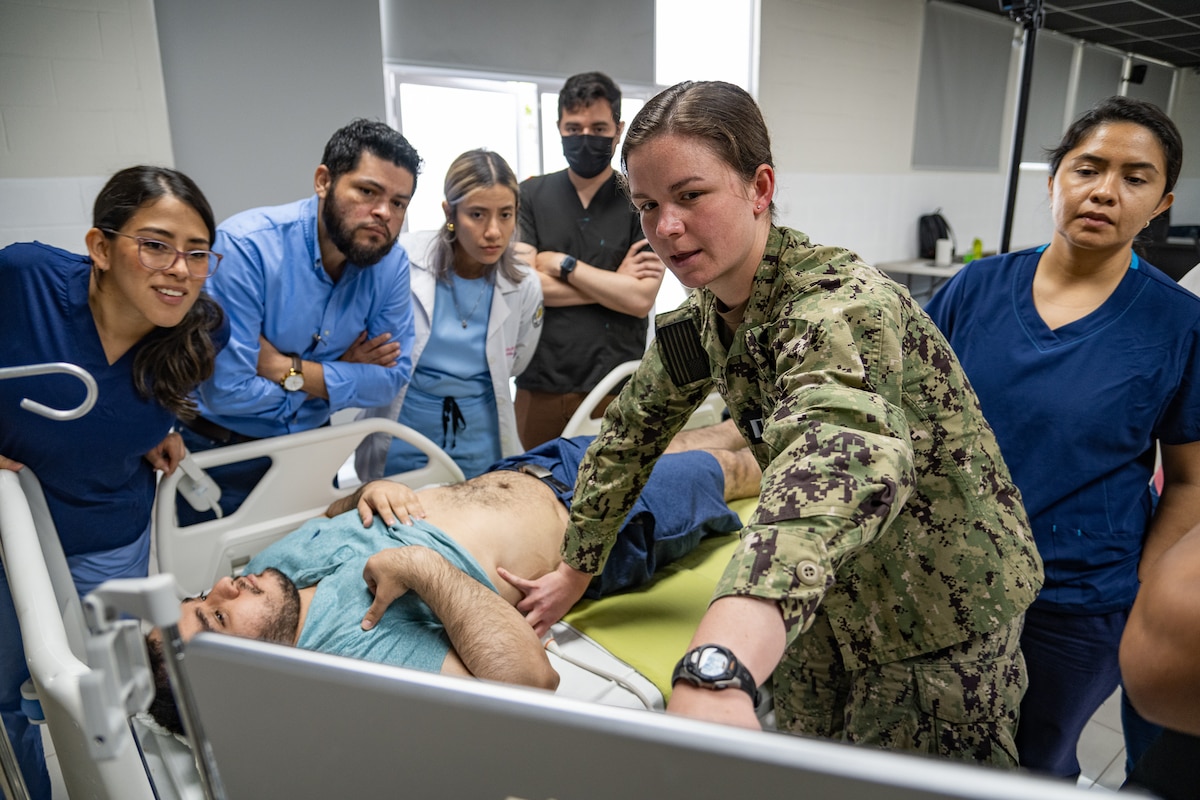 A woman in U.S. military uniform points at a screen while applying ultrasound gel to a man's abdomen as he lays in a hospital bed surrounded by men and women in civilian clothes.