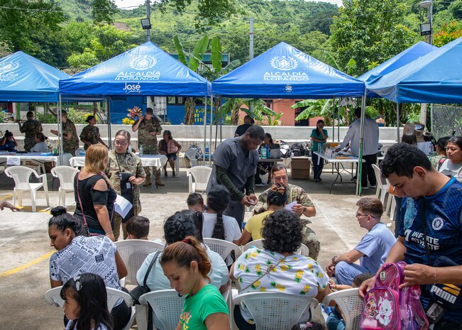 A group of people in a basketball court with tents in the background receive medical care by U.S. servicemembers.