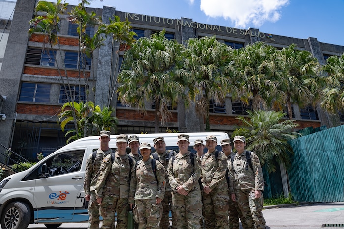 A group of U.S. service members stands in front of a building titled "INSTITUTO NACIONAL DE SALUD"