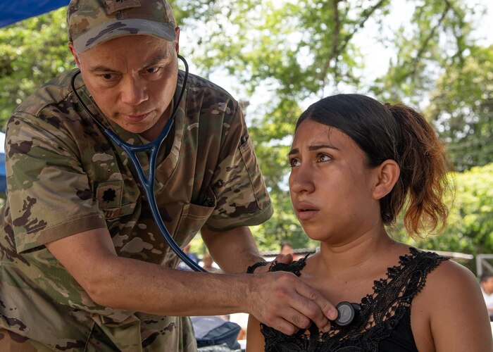 A man in U.S. military uniform holds a stethoscope to a woman's chest.