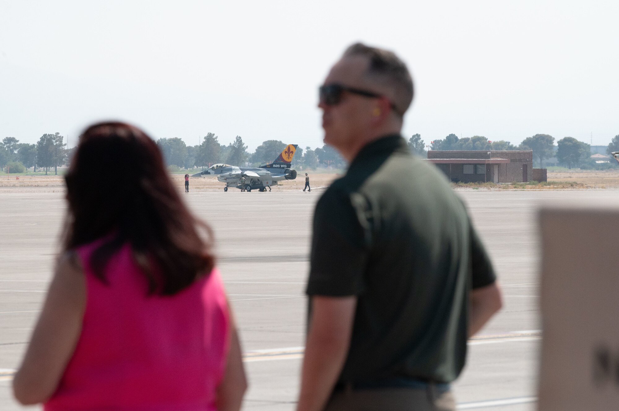 Two onlookers watch an F-16 fighter jet as it taxis on the flight line.