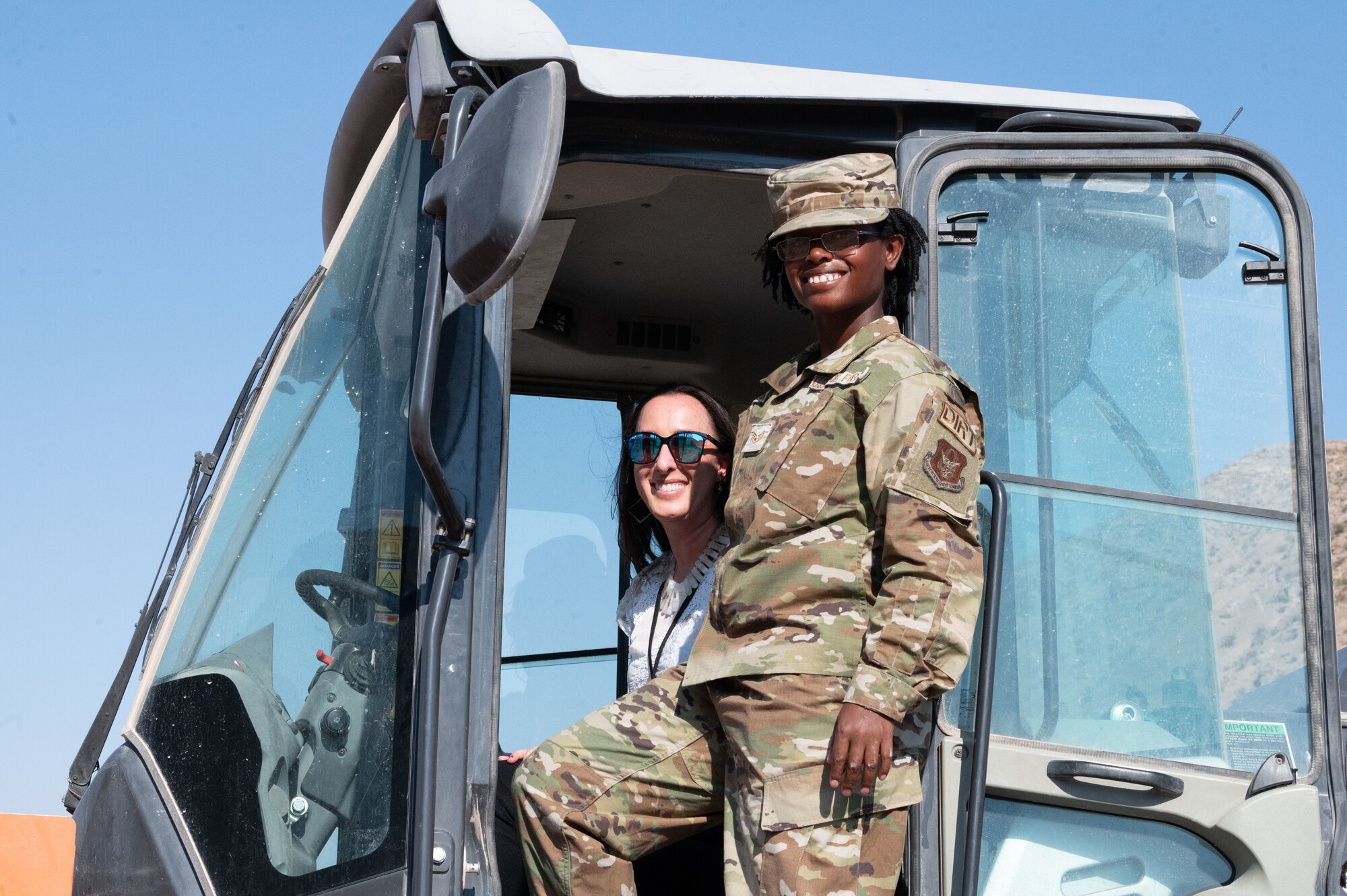 A woman in uniform stands in the doorway of a heavy equipment vehicle as another women sitting behind the control smiles at the camera.