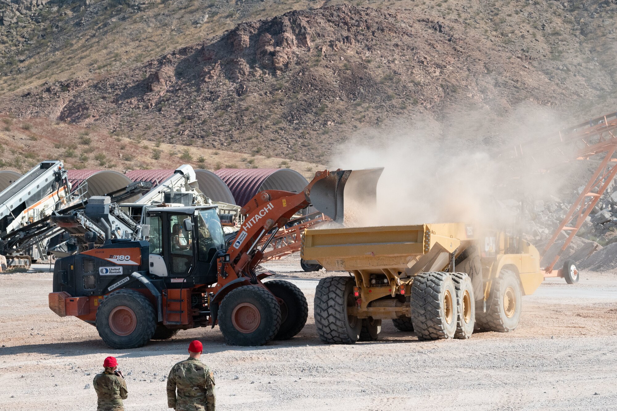 Two heavy machinery vehicles in a dirt lot as one uses its shovel to pour dirt into the bed of another.