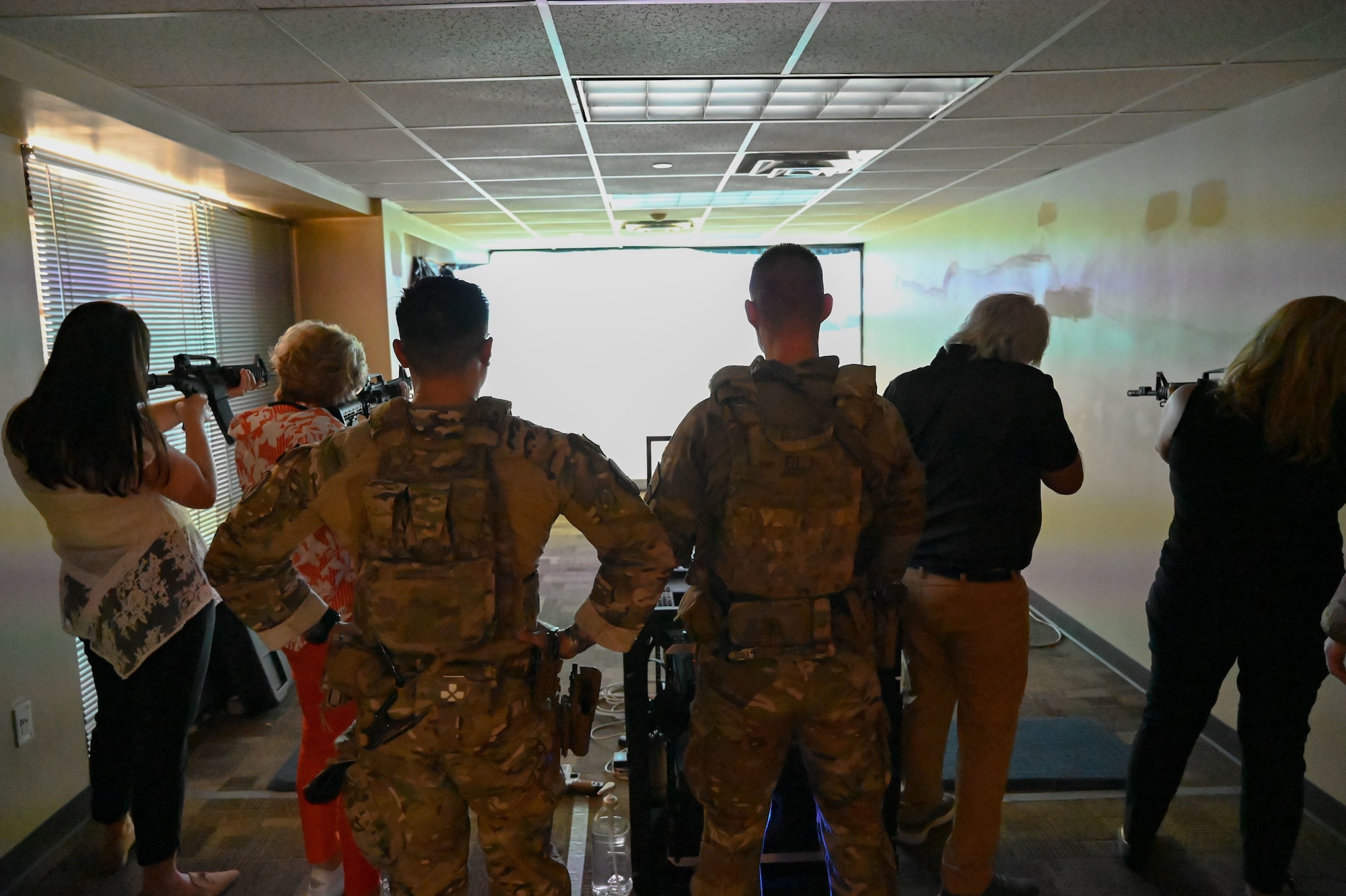 Military members look on as honorary commanders point simulation training weapons at a weapons qualification video simulation screen.