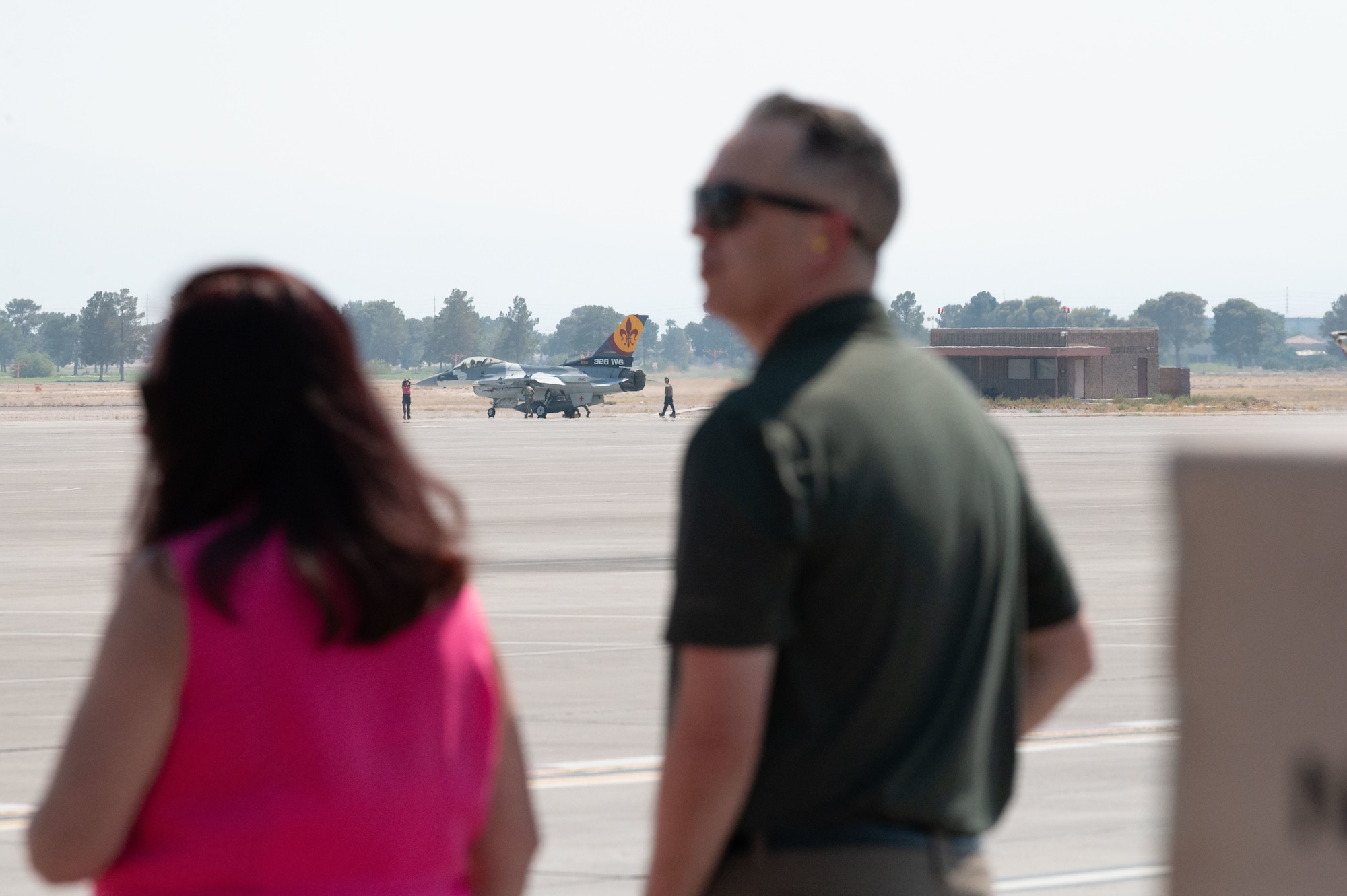 Two onlookers watch an F-16 fighter jet as it taxis on the flight line.