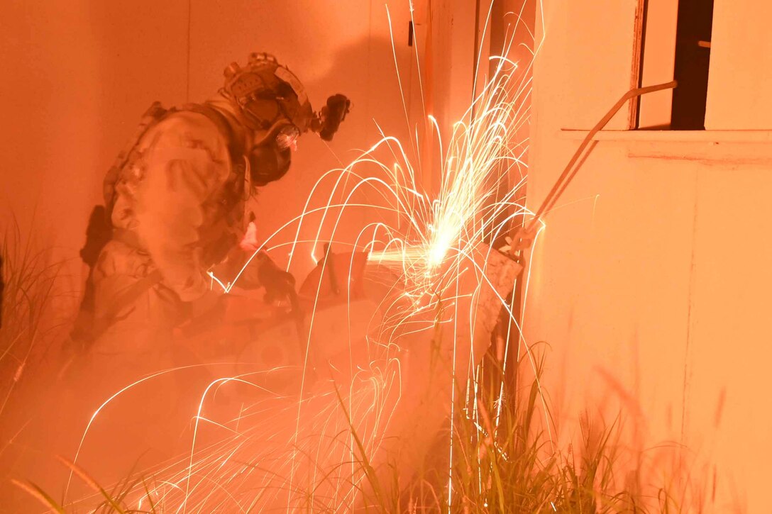 An airman wearing a helmet and goggles uses a saw to cut through a barrier creating sparks as outdoor lighting creates an orange glow.