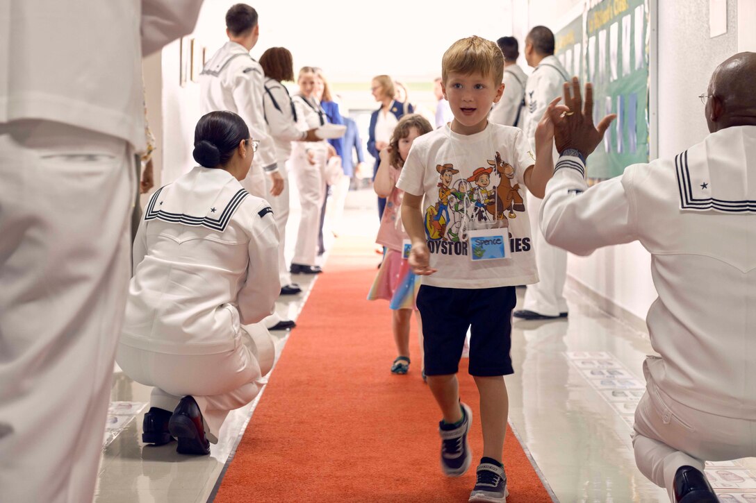 Students walk along an orangish-red carpet toward the camera as sailors give high-fives while kneeling and standing on each side of them.