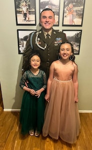 Soldier in dress uniform poses with two young girls in formal dresses