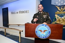 U.S. Army Soldier in dress uniform poses behind a podium with the Department of Defense crest on it in the Hall of Heroes.