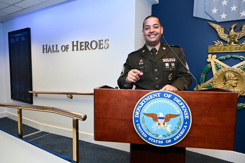 U.S. Army Soldier in dress uniform poses behind a podium with the Department of Defense crest on it in the Hall of Heroes.