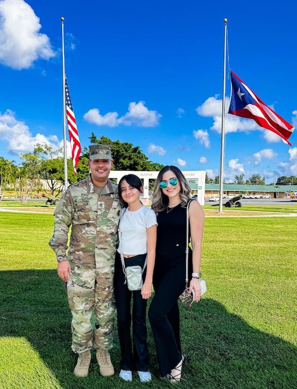 U.S. Army Soldier poses with wife and daughter in front of the American Flag and Puerto Rican Flag.